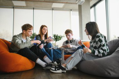 Dört genç yetişkin, modern ofis alanında puf koltuklarda oturuyor, kart oynuyor ve boş zamanlarının tadını çıkarıyorlar.