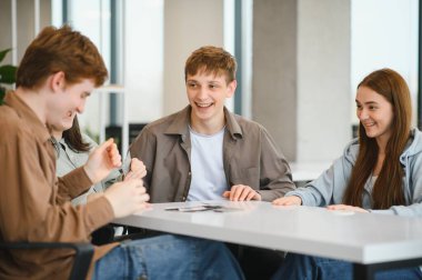 Dört neşeli üniversite öğrencisi beyaz bir masanın etrafında oturmuş oyun oynuyorlar ve ara verdiklerinde kahkahalarını paylaşıyorlar.