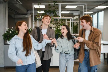 Sırt çantalı dört neşeli öğrenci birlikte eğleniyorlar. Parlak bir üniversite koridorunda gülüyorlar ve eğleniyorlar.