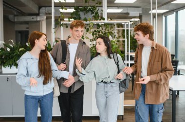 Dört mutlu öğrenci bir üniversite koridorunda birlikte yürüyüp konuşuyorlar, sırt çantaları taşıyorlar ve arkadaşça sohbet ediyorlar.