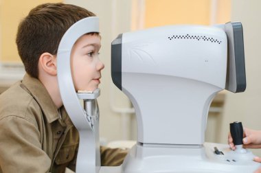Çocuk, modern bir klinikteki göz doktoruna yaptığı ziyarette göz muayenesi için tıbbi bir cihaz kullanıyor.