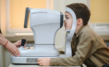 Bir göz doktoru bir klinikte detaylı bir göz muayenesi yaparken çocuk otomatik fraktör ile görme kontrolünden geçiyor.
