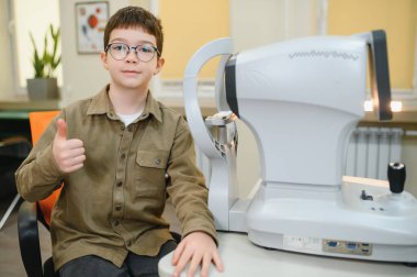 Gözlüklü bir çocuk göz hekiminde görme yeteneğini kontrol ettikten sonra baş parmağını gösteriyor.