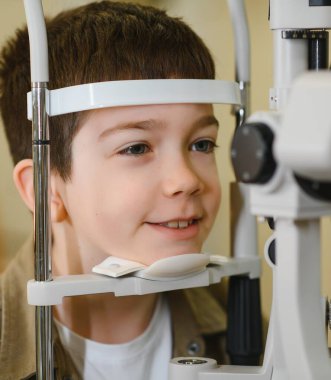 Gülümseyen çocuk modern bir göz hastalıkları kliniğinde bir phoropter ile görme kontrolünden geçiyor, tam bir göz muayenesinden geçiyor.