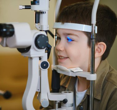 Bir göz hekiminde modern bir yarık lambasıyla biyomikroskop kullanarak görme kontrolünden geçen gülümseyen çocuk özel bir göz tedavisi görüyor.