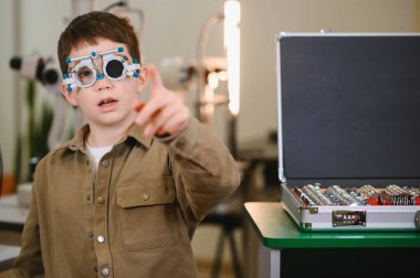 Deneme çerçevesi takan bir çocuk, bir göz sağlığı kliniğinde görme testi sırasında parmağıyla işaret ediyor, görme ve göz sağlığını değerlendiriyor.