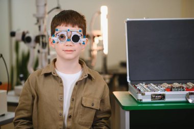 Deneme çerçevesi takan genç bir çocuk, göz doktorunun kliniğinde göz muayenesinden geçiyor, görme sağlığı ve bakımı üzerinde çalışıyor.