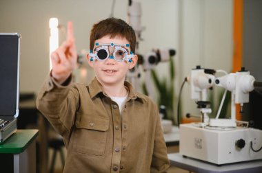 Optalmoloji kliniğinde görme testi sırasında parmağıyla işaret eden deneme çerçevesi takan genç bir çocuk.
