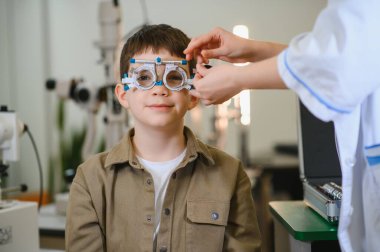 Oftalmolog, klinikte görüş kontrolleri sırasında gülümseyen çocuğu mahkeme çerçevesiyle inceliyor.