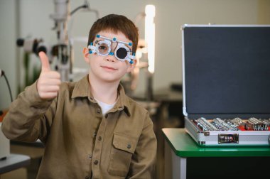 Deneme çerçevesi takan bir çocuk, göz muayenesinden geçerken başparmak kaldırıyor, mutluluk saçıyor.