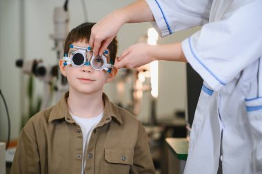 Oftalmolog, bir klinikte görme testi sırasında genç bir çocuğun yüzüne bir deneme çerçevesi yerleştiriyor. Görme yeteneğini dikkatlice inceliyor.