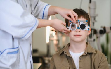 Oftalmolog, bir klinikteki görme testi sırasında bir çocuğun yüzündeki fotoropteri düzeltiyor ve çocukların göz bakımını gösteriyor.
