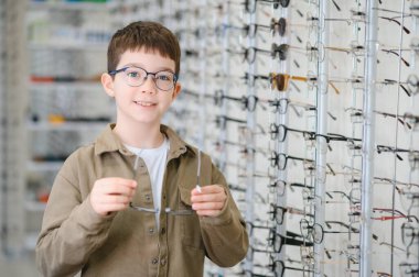 Optalmoloji kliniğinde gözlük takan gülümseyen çocuk, gözlükçülerin yardımıyla çerçeveler seçiyor, daha iyi görebilmek için yeni gözlüklerle mutlu.