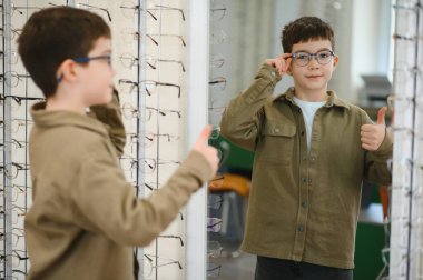 Bir optik mağazasında yeni gözlük deniyor, gülümsüyor ve başparmak kaldırıyor, şık gözlük seçiminden açıkça memnun.