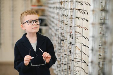 Gözlüklü çocuk optik mağazada yeni çerçeve seçiyor, görüş düzeltmesi için çeşitli seçenekler keşfediyor