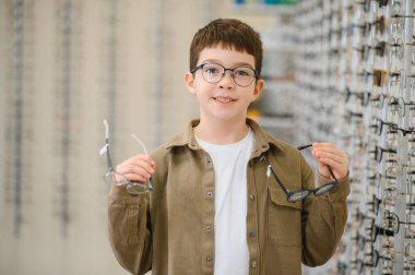 Gözlüklü gülümseyen çocuk iki çift çerçeve tutuyor, optik mağazada yeni gözlük seçiyor, oftalmoloji kliniği
