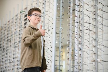 Yeni gözlük takan bir çocuk, gözlük seçiminden memnun, gözlükçü dükkanında baş parmak işareti yapıyor.