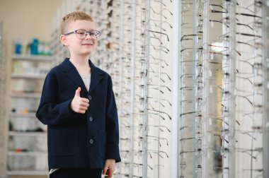 Yeni gözlük takan gülümseyen çocuk başparmağını kaldırıyor. Göz hekimliği seçiminden memnun.