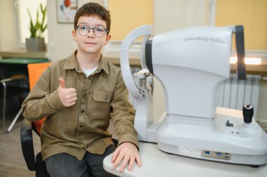 Gözlüklü genç, göz hekimliği kliniğinde otorefraktörle görme testinde baş parmak gösteriyor.