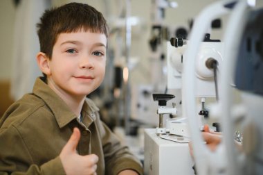 Göz doktorunun muayenesinde baş parmak gösteren genç bir çocuk. Arka planda göz doktoru ekipmanları var.