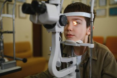Klinikte biyomikroskop kullanarak gözlerini göz doktoruna muayene ettiren genç bir çocuk.