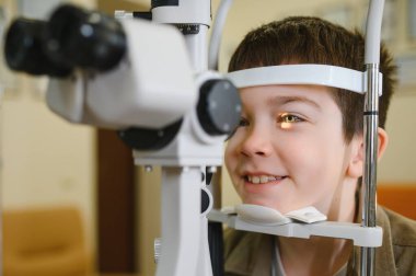 Bir göz hekimi kliniğinde gelişmiş ekipmanlarla görme kontrolünden geçen gülümseyen çocuk, bir sağlık uzmanından uzman bakımı alıyor.
