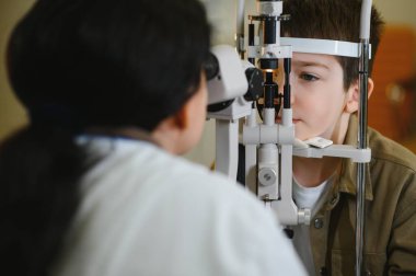 Modern bir klinikte çalışan bayan bir göz doktoru tarafından desteklenen kesik lamba biyomikroskobu ile göz muayenesinden geçen genç bir çocuk.