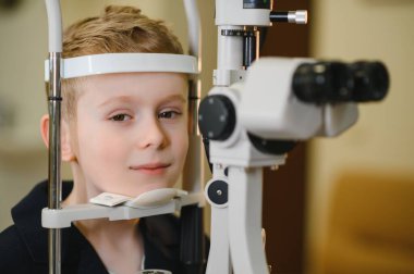 Göz muayenesinden geçen genç bir çocuk göz hekimliği kliniğinde phoropter kullanıyor, görme ve teşhis konularını vurguluyor.