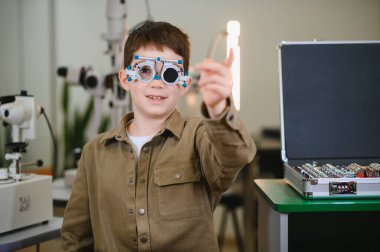 Göz doktorunun muayenehanesinde görme testinde parmağıyla işaret eden deneme çerçevesi takan genç bir çocuk.