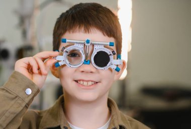 Göz muayenesi sırasında bir göz doktorunun muayenehanesinde deneme çerçevesi takan gülümseyen çocuk pediatrik göz bakımının önemini gözler önüne seriyor.
