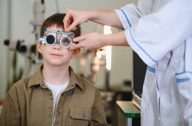 Oftalmolog, klinikteki görme testi sırasında genç bir çocuğun yüzündeki bir deneme çerçevesini düzeltiyor. Potansiyel sorunlara karşı görme yeteneğini kontrol ediyor.