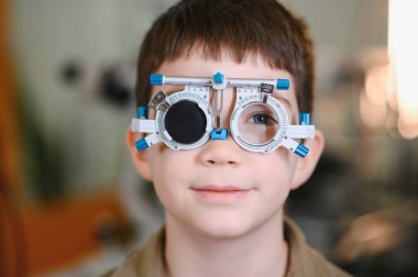 Deneme çerçevesi takan genç bir çocuk, bir göz doktorunun muayenehanesinde görme muayenesinden geçiyor, kontrol sırasında gülümsüyor.