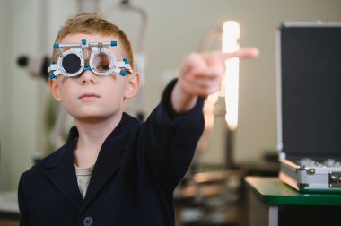 Deneme çerçevesi takan bir çocuk, göz muayenesi sırasında parmağıyla işaret ederek bir göz doktorunda görme kontrolüne giriyor.