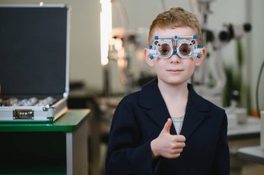 Bir göz doktorunun muayenehanesinde göz muayenesi sırasında mutluluk ve güven saçan, deneme çerçevesi takan ve onay veren bir çocuk.