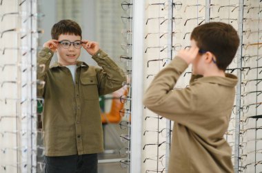 Çocuk bir optik mağazada aynanın önünde yeni gözlük deniyor. Yeni görünüşünü farklı çerçevelerle kontrol ediyor.