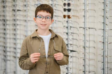 Gülümseyen çocuk gözlük takıyor ve bir optik mağazada bir çift çerçeve tutuyor. Gelişmiş görüş için yeni gözlük seçiyor.