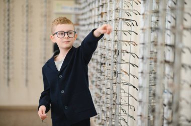 Gözlük takan genç bir çocuk bir optik mağazadaki gözlük ekranını işaret ederek gelişmiş görüş için yeni çerçeveler seçiyor.
