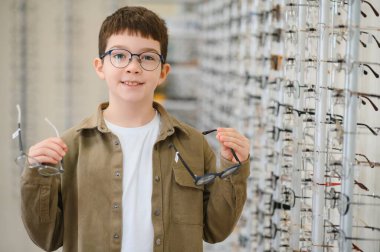 Gözlüklü genç, bir optik mağazada farklı çerçeveleri karşılaştırırken gülümsüyor ve çocuklar için görme bakımının önemini vurguluyor.