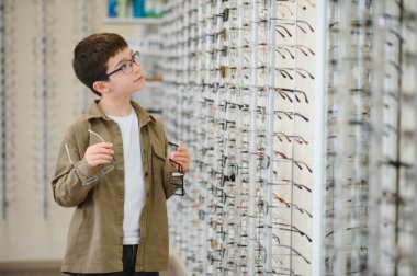 İki gözlük tutan çocuk optik mağazada yeni gözlük, göz hekimliği ve çocuk vizyon düzeltmesi seçiyor.