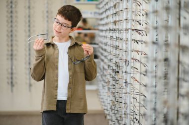 Çocuk iki gözlük çerçevesini karşılaştırıyor, bir optik mağazada karar veriyor, çocuklar için vizyon bakımının önemini vurguluyor.