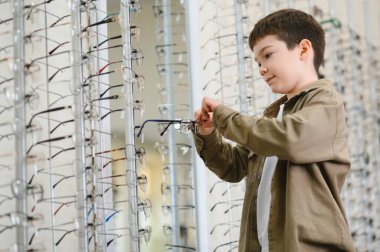 Çocuk, optik mağazadaki vitrinlerden gözlük seçerken, çocuklar için görüş bakımının önemini gösteriyor.