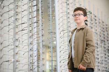 Yeni gözlük takan çocuk optik mağazada gözlük seçiyor, göz doktoru kliniğinde gülümseyen çocuk farklı çerçeveler deniyor.