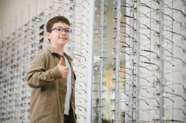 Optik mağazasında, göz hekimliği kliniğinde, çocuk vizyonu düzeltmesinde ve göz bakımı konseptinde baş parmak hareketi gösteren yeni gözlük takan mutlu çocuk.