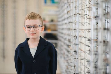 Yeni gözlük takan ve kendine güvenen bir çocuk canlı bir gözlük vitrininin yanında dikilirken kendine güvenen bir şekilde gülümsüyor.