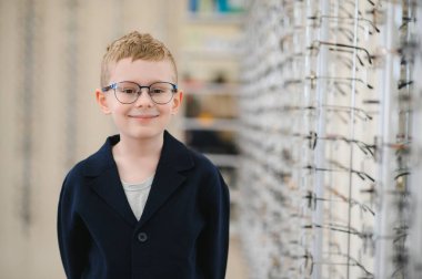 Göz hekimliği kliniğinde yeni gözlük takmış gülümseyen genç bir çocuk, bir gözlük çerçevesinin yanında duruyor.