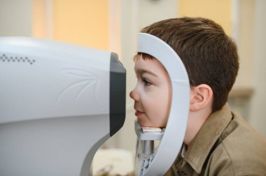 Bir sağlık kliniğinde göz muayenesi sırasında modern bir oto-fraktörle meşgul olan çocuk hasta, görme kontrolü ve bakımı üzerine odaklanıyor.