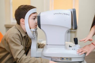 Çocuk, modern oftalmoloji kliniğinde otomatik fraktör kullanılarak görsel keskinlik muayenesinden geçiyor. Göz doktoru, çocuğun görme yetisini inceliyor.