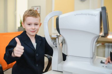 Görme testi sırasında göz hekimliği kliniğindeki modern ekipmanlarla başparmak kaldıran genç bir çocuk, deneyimi hakkında olumlu geri bildirim gösteriyor.