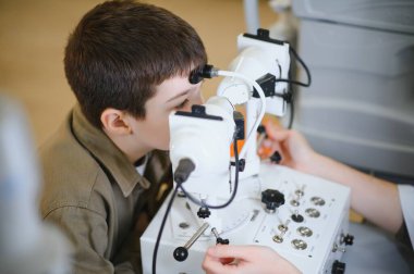 Oftalmolog, klinikte genç bir çocuğun görme yeteneğini inceliyor. Doğru teşhis ve tedavi için gelişmiş tıbbi gereçler kullanıyor.