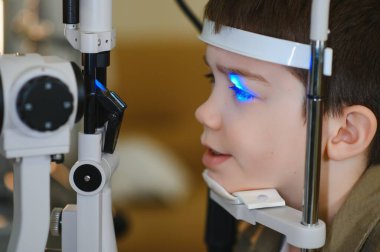 Özel ekipmanlarla vizyon kontrolünden geçen genç bir çocuk, modern bir klinikte bir göz doktorunun yardımıyla, doğru teşhis ve tedaviyi garanti altına alıyor.
